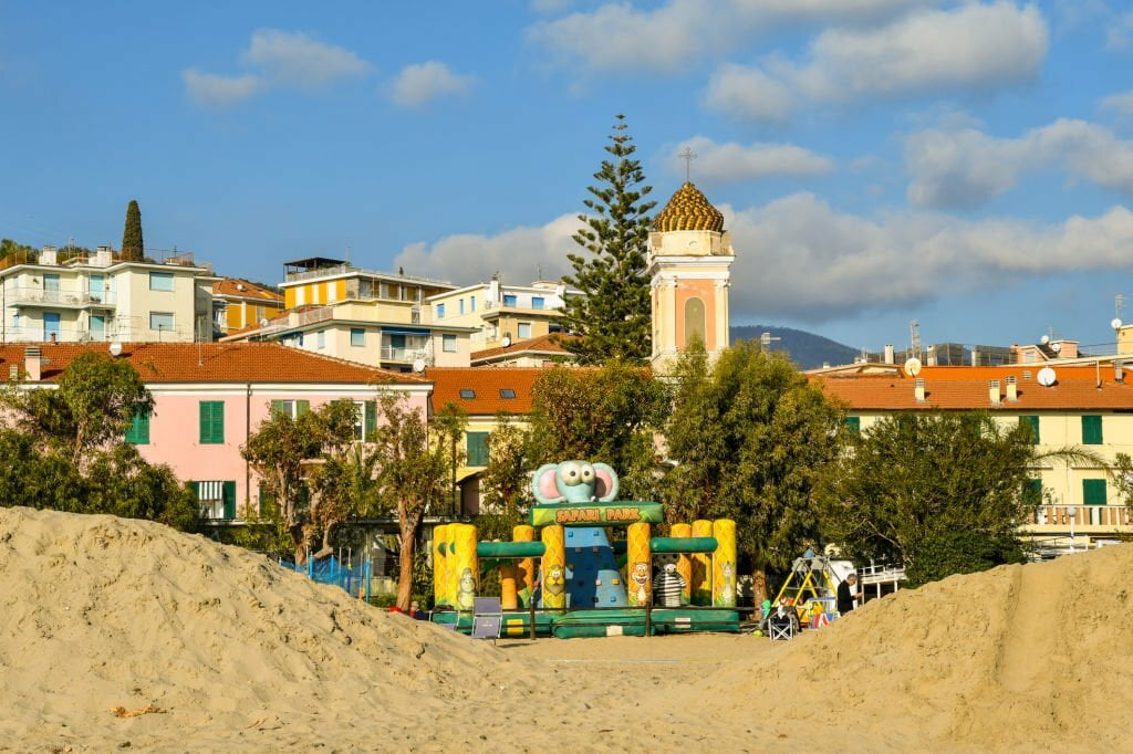 Une scène de plage avec un château gonflable et une aire de jeux au premier plan, entourée de dunes de sable. Derrière, on a une vue pittoresque de bâtiments et de maisons aux façades colorées, d'arbres et d'une église avec un clocher sur un ciel bleu avec des nuages épars.