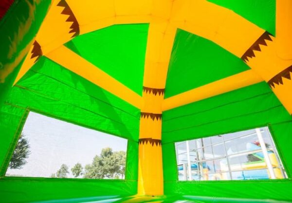 L’image montre la vue intérieure d’un Château Maxifun Super Crocodile coloré, vert et jaune. Les murs comportent de grandes fenêtres claires à travers lesquelles les arbres et certains équipements de jeux sont visibles. Le toit présente un motif entrecroisé avec des détails pointus et marron.