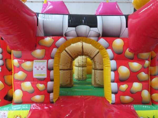 Une maison gonflable colorée ressemblant à un château avec un extérieur tacheté de rouge et de jaune, des tours cylindriques au sommet et une entrée voûtée. Un petit panneau avec les consignes de sécurité est visible à côté de l'entrée, permettant à tous de profiter en toute sécurité de ces Parcours de jeu : L'attaque du Dragon 25 m.