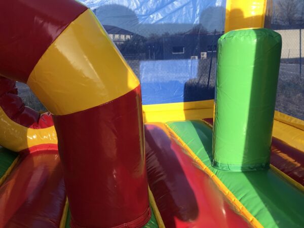 L'image montre l'intérieur coloré d'un jeu gonflable : Primairo avec des sections rouges, jaunes, vertes et bleues. Deux grandes colonnes gonflables cylindriques, une rouge et jaune et une verte, se tiennent debout. Un flanc en filet et un fond de montagnes sont visibles.