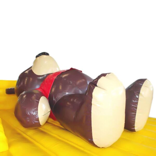 Un grand CHÂTEAU GONFLABLE NOUNOURS repose sur le dos sur une surface gonflable jaune. L'ours est principalement brun avec un ventre et un museau de couleur crème et porte un gilet rouge. Le fond est blanc et uni.
